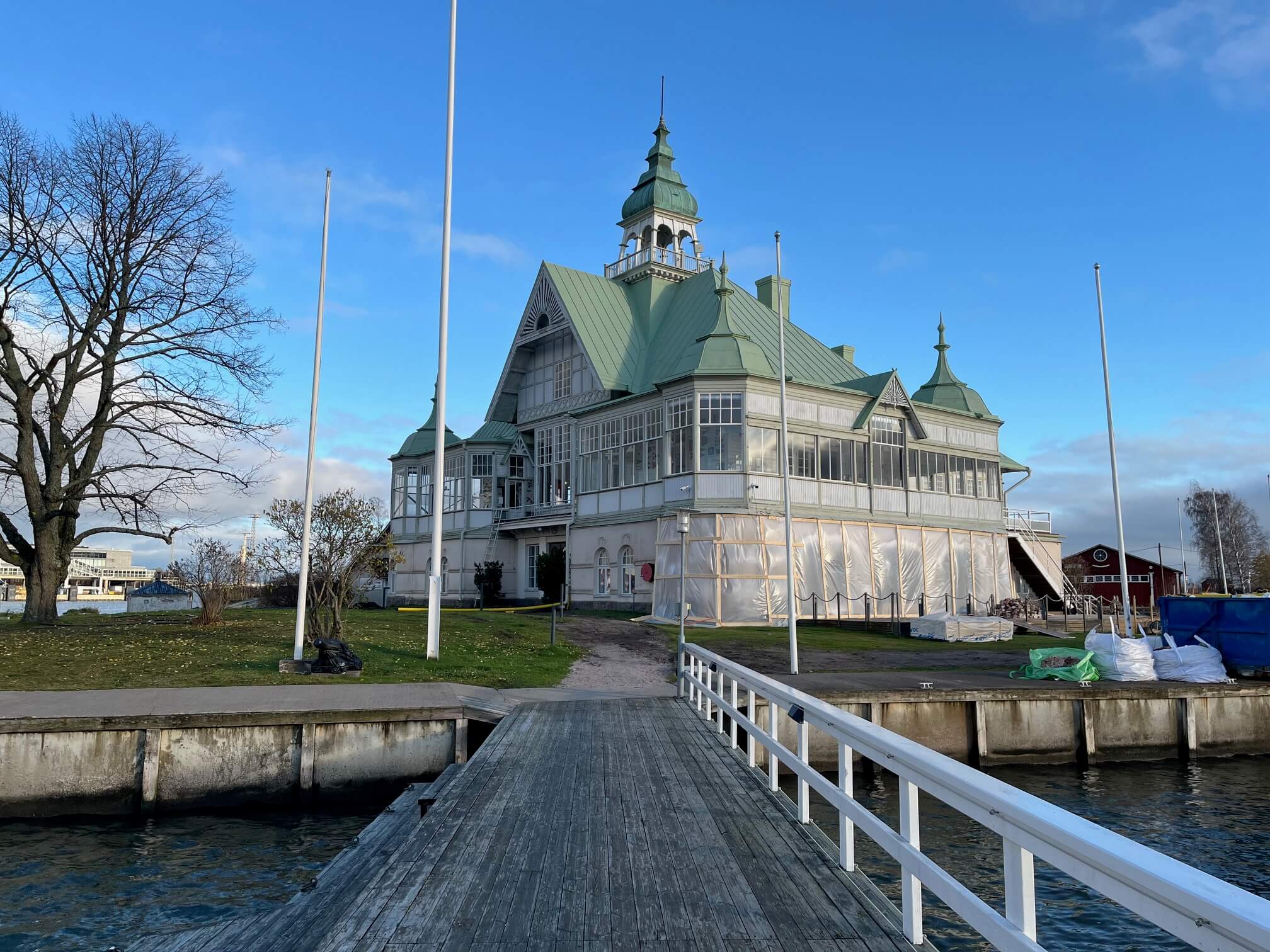 Kuvassa on puinen laituri, jonka päässä on suuri, vaalea rakennus vihreällä katolla ja tornilla. Taustalla näkyy sininen taivas.