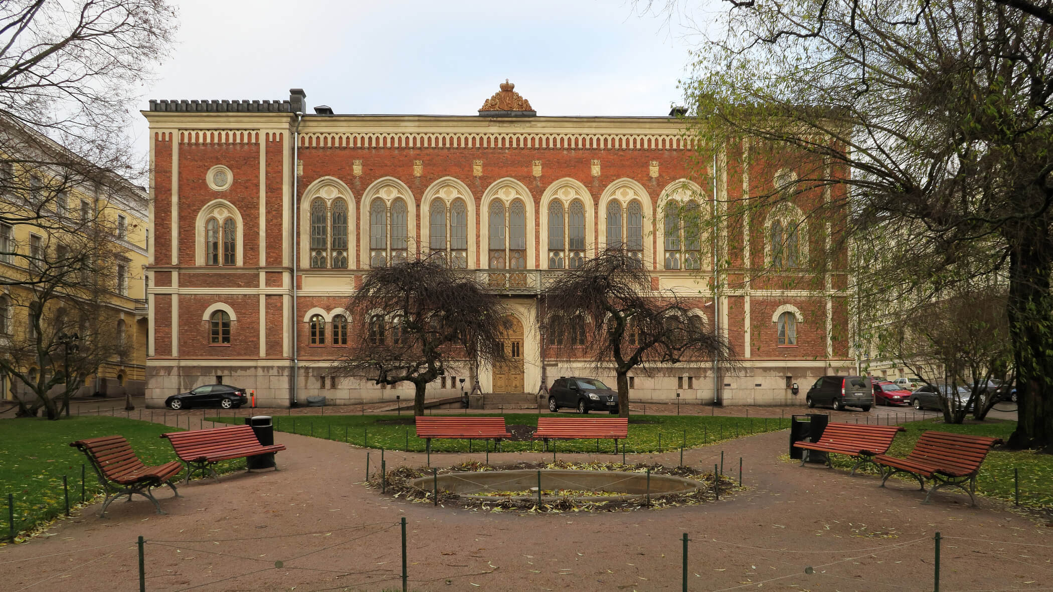 Punatiilinen rakennus puiston keskellä, jossa on penkkejä ja kuivia puita. Taustalla näkyy autoja ja harmaa taivas.